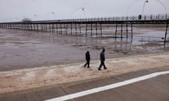 two people walk along promenade by Southport pier, Merseyside, at low tide on a grey, wintry day