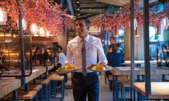 Sunak in a shirt and tie, with a name badge, carrying two plates of food to a table in an illuminated outdoor dining area.