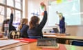 Pupils in a classroom raise their hands as a teacher points to writing on a whiteboard