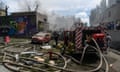 A fire engine with hoses running from it, as smoke rises from the rubble of a building in the background. To the left is a brightly coloured mural on a wall