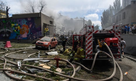 A fire engine with hoses running from it, as smoke rises from the rubble of a building in the background. To the left is a brightly coloured mural on a wall