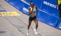 Hellen Obiri, hair pulled back and wearing a tank top, running shorts and On trainers, grins as she runs past a barrier reading "Bank of America" at the Boston Marathon