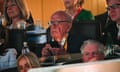 A very old white man with a mostly bald head and dark-rimmed glasses, wearing an orange lanyard and suit without a tie, looks up from his phone and appears to listen to something likely very far away from where he is sitting.