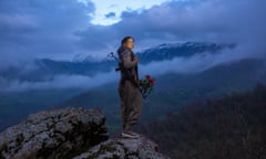 A woman holding a bunch of red flowers stands on a mountain top against a dramatic cloudy sky