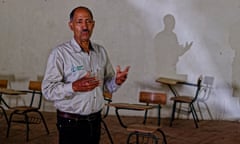 A man in a shirt and jeans standing in a classroom with empty desks