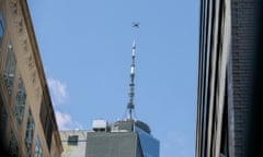 Shot from the street facing a blue sky, the photo is framed on both sides by the tops of skyscrapers, with an X-shaped drone seeming to hover above an telecommunications tower atop a skyscraper in the middle.