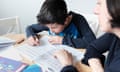 A female private tutor sits beside a young boy as he writes in a textbook