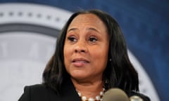 A closeup of a middle-aged Black woman, with straight black shoulder-length hair parted in the middle, a large white pearl necklace, and a black suit, speaking into a microphone before a government crest.