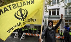 Middle-aged Sikh man with long white beard and black turban raises left fist while holding large yellow flag with black writing that says KHALISTAN and has a symbol and other writing on it.