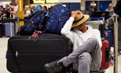 a delayed passenger snoozing at Manchester airport: he wears a straw hat pulled down over his face, a white hoodie, grey jeans and black trainers as he rests with his legs crossed, next to a huge black suitcase and two blue rucksacks. Other baggage and people can be seen in the background under bright, harsh lighting.