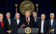 A line of older white men, all with gray hair, dark suit jackets, and no ties, stands behind an older white man with poofy hair speaking at a lectern, but he is wearing a tie.