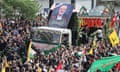 A truck decorated in flags, posters of the deceased and tassles, transports two coffins and is surrounded by a crowd of people who are carrying Iranian and Palestinian flags.