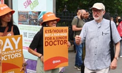 Jeremy Corbyn meets striking junior doctors outside St Thomas's hospital: he is wearing a loose short-sleeved cotton shirt and baseball cap and is seen talking to two young, female doctors who wear orange hats with the logo of the British Medical Association and carry placards reading 'pay restoration for doctors' and '£15 an hour is not a fair wage for a junior doctor'.