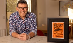 A man in glasses and a collared shirt leaning on a counter. Next to him is a framed handbill advertising a gig from The Birthday Party in 1983.