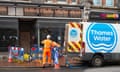 Two men wearing orange hi-vis overalls, one in a white hard hat, are seen around a hole in the pavement with lengths of blue piping; there are sandbags, barriers and hazard cones around the hole, and a Thames Water van bearing the company's logo parked by. A boarded-up shopfront without signage is seen behind the hole, and a takeaway noodle bar next to that.