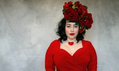 Bink wearing a large velvet rose fascinator, heart necklace and low-cut dress, slightly smiling