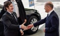 Two men in suits shake hands in front of a black car