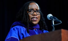 A middle-aged Black woman with long black hair, black-framed glasses, and a bright blue graduation robe speaks into a microphone at a lectern.