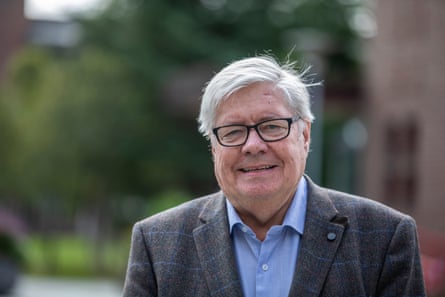 Smiling older gentleman with side-parted grey hair, wearing open-neck shirt and grey blazer.