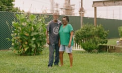 A middle-aged man and woman stand in a neatly groomed yard while an oil refinery looms in the background.