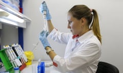 White woman with blond ponytail, white smock and powder blue gloves holds something test-tube-like in a lab.