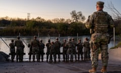 Soldiers stand guard in a line in front of a fenced area that gives access to the river beyond.