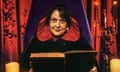 Kathy Burke sitting on a purple throne-like chair wearing a black cloak with a red-lined hood and holding an big black book and looking mischievous
