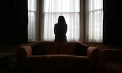 A woman stands alone in the bay window of a house.