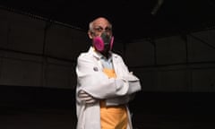 An elderly man  wearing a facemask, white lab coat and yellow apron looks at the camera