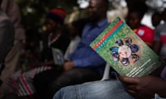 Africans sit listening to someone speaking while someone in the foreground holds a pamphlet with information about a medical trial