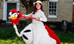 Rose, grinning, standing on stilts and holding reins to the head of an ostrich in an ostrich costume