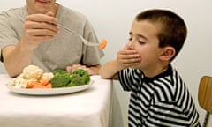 A boy sitting at a table with a plate full of vegetables and his hand over his mouth