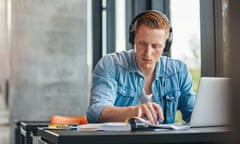 University student working on academic assignment<br>Portrait of young student wearing headphones sitting at the table in library and reading book. University student finding information for his academic assignment.