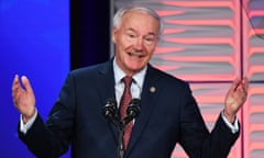 Older white man with white hair holding both hands up as he speaks into two microphones at a lectern on a stage, with electric blue and red lights behind him.