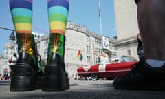 A photo taken from street level, between the ankles of someone wearing rainbow socks on the left and another set of ankles on the right. In the center, in the street, is a bright red convertible decorated with rainbow flags, with a blond woman in the back waving to crowds.