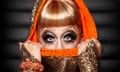 Close-up of Bianca Del Rio's elaborately made-up face, half-covered with an orange and gold hood, which she pulls up over her mouth with her hands.