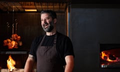 Chef Ben Williamson, dressed in a brown apron and black T-shirt, laughs in his restaurant kitchen while a wood-fire grills burns in the background