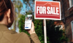 Rear view of woman using smartphone while looking at a for sale sign on a house