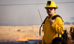 A man wearing a yellow button-down and yellow hard hat stands in front of burning grass