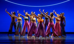 a group of 12 dancers in colourful costumes, against a deep blue backdrop and beneath a neon semicircle, each with their right hand raised, palm flat