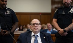 Man in suit and glasses sits flanked between security guards in court