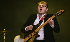 Alex Moore of the Lathums with a fringe, glasses and jacket over a shirt singing and playing guitar