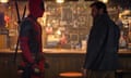 Two men stand at a bar, one wearing a red and black suit and the other wearing a dark jacket