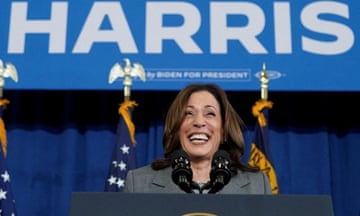A woman wearing a gray blazer smiles as she stands at a podium in front of a blue sign reading 'Harris'.