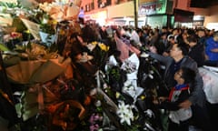 Crowds of people stand looking at a huge pile of flower tributes.