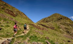 Two people hike up a mountain path. The area around them is grassy and there is a clear blue sky.