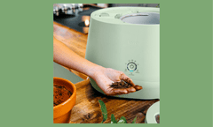 Image of light-skinned hand holding dirt, next to a white plastic machine on a wooden counter next to a terra cotta pot.