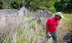 In an overgrown field, a middle-aged Black man wearing a red T-shirt and baseball cap walks away from the fallen-down stone walls of a building.