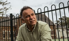 John Pesutto is leaning on a waist-high fence with a higher fence, a church and a tennis court behind him