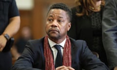 A Black man with trim hair a bit long on top, in black suit with red scarf, sits with hands crossed on table in front of him, looking serious and/or anxious toward someone on his right.